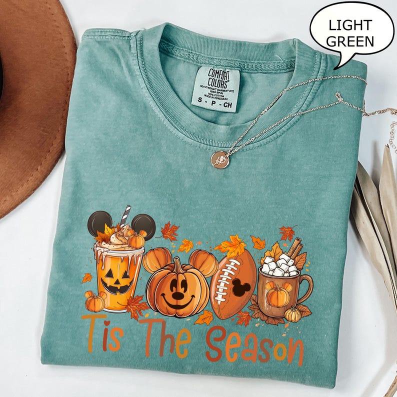 May include: Light green t-shirt with a fall-themed graphic. The design includes a pumpkin spice latte, Mickey Mouse pumpkins, a football, and a mug of hot cocoa. The text "Tis The Season" is printed in orange.