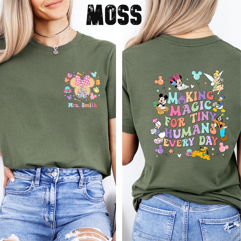 May include: Moss green t-shirt with a colourful graphic on the front featuring a Minnie Mouse design and the text "Mrs. Smith". The back of the shirt has a graphic with Disney characters and the text "Making Magic for Tiny Humans Every Day".