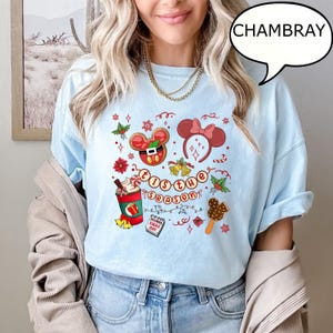May include: Pale blue t-shirt featuring a Christmas-themed design. The graphic includes Mickey Mouse in a Santa suit, Minnie Mouse ears, a festive drink, and the text "'Tis the Season". The shirt is made of chambray fabric.