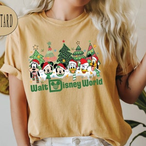 May include: A mustard yellow t-shirt with a Christmas-themed design featuring Mickey Mouse, Minnie Mouse, Donald Duck, Goofy, and Pluto. The design includes Christmas trees, snow, and the text "Walt Disney World".