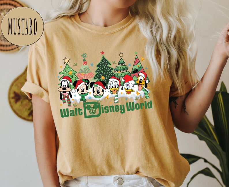 May include: A mustard yellow t-shirt with a Christmas-themed design featuring Mickey Mouse, Minnie Mouse, Donald Duck, Goofy, and Pluto. The design includes Christmas trees, snow, and the text "Walt Disney World".