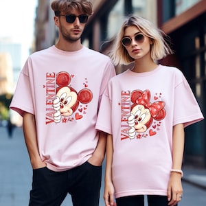 Puede incluir: Dos personas con camisetas rosas a juego. Cada camiseta presenta un dise&ntilde;o de personaje de dibujos animados con la palabra "VALENTINE" impresa verticalmente. El dise&ntilde;o incluye un rat&oacute;n de dibujos animados con orejas rojas y corazones.