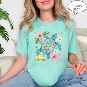 May include: A light turquoise t-shirt featuring a watercolor sea turtle design. The turtle has a teal shell with pink and yellow floral accents. The shirt also includes the text "ISLAND REEF" in a speech bubble.