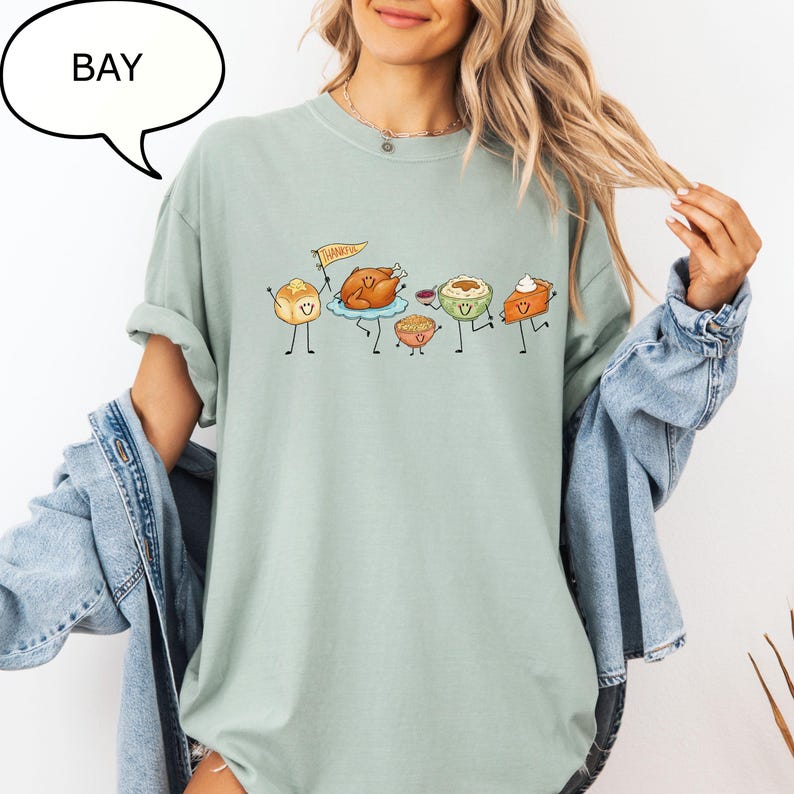 Pu&ograve; includere: Una t-shirt girocollo color verde salvia con una grafica di cibi del Ringraziamento antropomorfi, tra cui un tacchino, un panino con una bandierina con la scritta THANKFUL, pur&egrave; di patate, ripieno e una fetta di torta di zucca.
