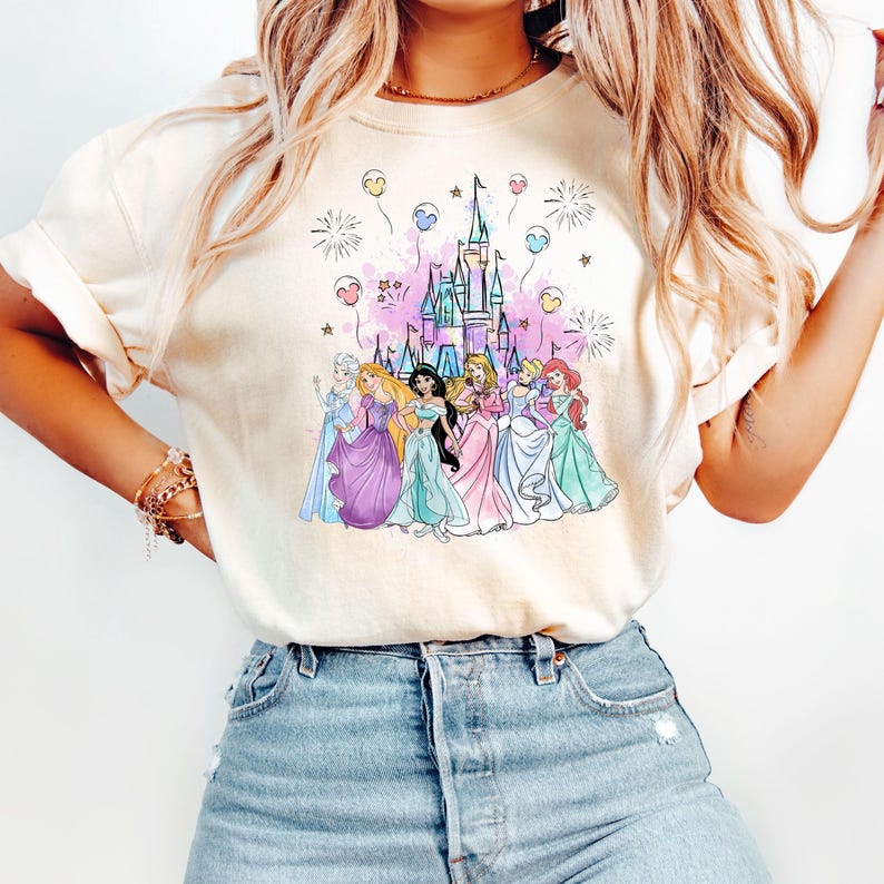 Puede incluir: Una camiseta blanca con una ilustraci&oacute;n colorida de princesas Disney de pie frente al castillo de Cenicienta. El castillo est&aacute; rodeado de fuegos artificiales y globos rosas y azules.