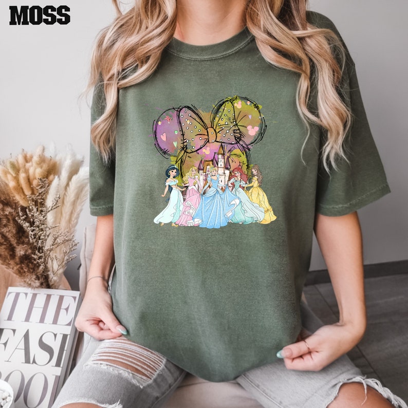 Puede incluir: Camiseta verde oliva con un dise&ntilde;o caprichoso de princesas de Disney, un castillo y un gran lazo con orejas de rat&oacute;n. La palabra "MOSS" est&aacute; impresa en la parte superior izquierda.