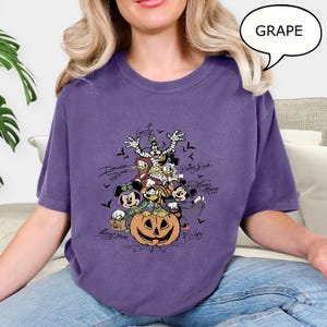 May include: A purple t-shirt with a graphic of Disney characters dressed for Halloween. The design includes Mickey Mouse, Minnie Mouse, Donald Duck, Goofy, and Pluto, with a carved pumpkin and bats. The shirt is labeled "Grape".