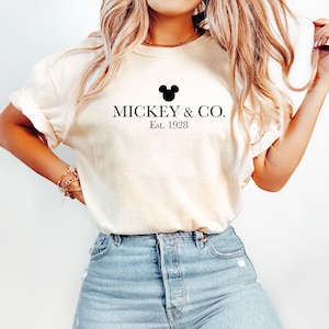 Puede incluir: Una camiseta blanca con una cabeza de Mickey Mouse negra y el texto "MICKEY & CO. Est. 1928" impreso en la parte delantera.