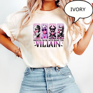 May include: Ivory-colored t-shirt featuring a graphic of four cartoon villains blowing bubblegum bubbles. The text reads "Sweetie I'm ok with being the Villain" in purple, with bat silhouettes. The shirt is a casual, everyday wear item.