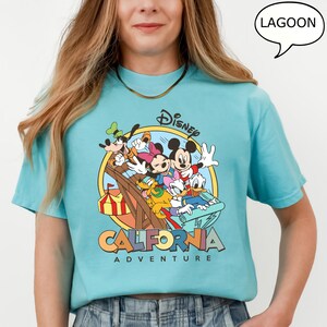 Puede incluir: Una camiseta azul claro con un estampado gr&aacute;fico de Mickey Mouse y sus amigos montando una monta&ntilde;a rusa. El gr&aacute;fico incluye el texto "Disney California Adventure" y un bocadillo que dice "Lagoon".