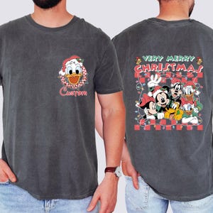 May include: Dark gray t-shirt featuring a Christmas-themed design. The front displays Donald Duck in a Santa hat with the word "Custom" below. The back showcases Mickey, Minnie, Goofy, Donald, and Pluto with "Very Merry Christmas" text.