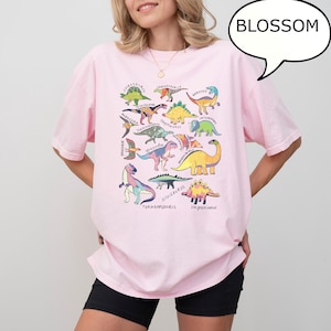 May include: A light pink t-shirt featuring a colorful dinosaur graphic. The design includes various dinosaur illustrations with their names, such as Tyrannosaurus and Stegosaurus. A speech bubble with the word "BLOSSOM" is on the top right.