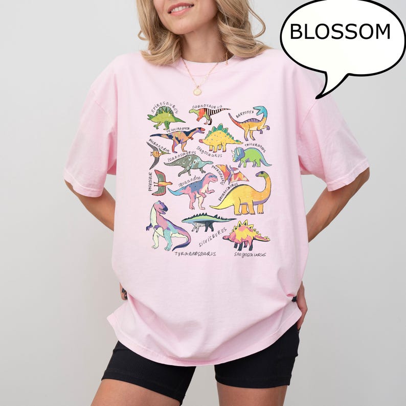 May include: A light pink t-shirt featuring a colorful dinosaur graphic. The design includes various dinosaur illustrations with their names, such as Tyrannosaurus and Stegosaurus. A speech bubble with the word "BLOSSOM" is on the top right.