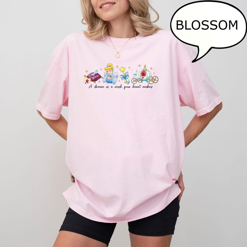 K&ouml;nnte beinhalten: Ein hellrosa T-Shirt mit einer bunten Grafik von Cinderella, einer Kutsche und dem Text "A dream is a wish your heart makes." Das Wort "BLOSSOM" befindet sich in einer Sprechblase. Das T-Shirt wird von einer Person getragen.