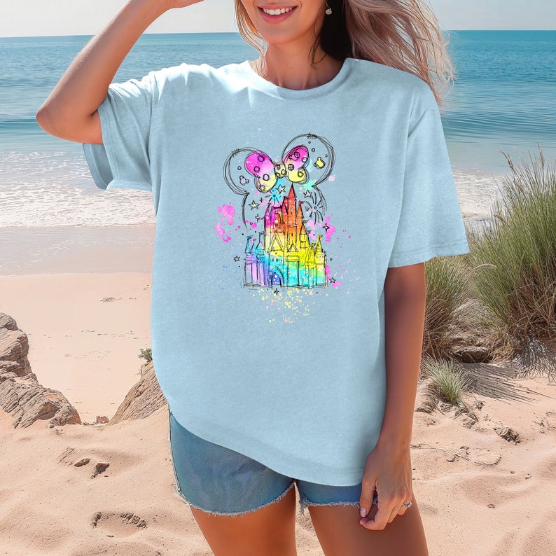 Puede incluir: Una camiseta azul claro con un gr&aacute;fico colorido de un castillo con un lazo en la parte superior. El castillo est&aacute; rodeado de estrellas y brillos.