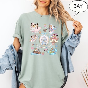 Puede incluir: Camiseta verde salvia con un collage de dibujos animados y el texto "BEST DAY EVER". El dise&ntilde;o incluye un castillo, Mickey y Minnie Mouse, el Pato Donald y otros personajes. La camiseta tiene mangas cortas y un corte holgado.