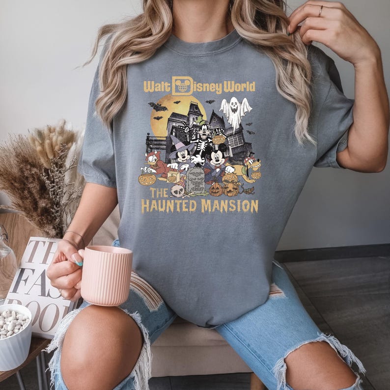 Puede incluir: Una camiseta gris con un gr&aacute;fico de Mickey Mouse y sus amigos frente a la Mansi&oacute;n Encantada en Walt Disney World. El texto "Walt Disney World" y "The Haunted Mansion" est&aacute; impreso en la camiseta.