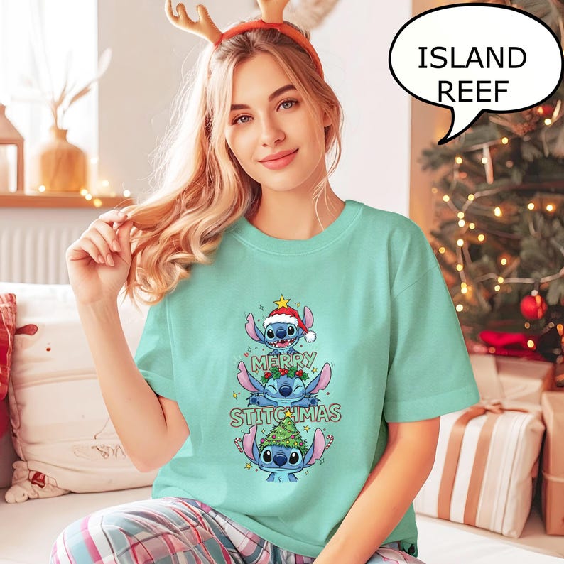 May include: A light teal t-shirt featuring a stacked cartoon character design with the text "MERRY STITCHMAS". The character is wearing a Santa hat and a Christmas tree. A speech bubble says "ISLAND REEF".