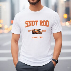 Puede incluir: Camiseta blanca con un gr&aacute;fico naranja de un coche de dibujos animados con el texto "SNOT ROD EST 1968 DISNEY CAR"