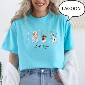 K&ouml;nnte beinhalten: Hellblaues T-Shirt mit einer Grafik von Figuren aus dem Film Frozen, darunter Elsa, Olaf und Sven, mit dem Text "Let it go". Das Wort "Lagoon" befindet sich in einer Sprechblase.