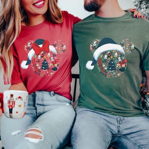 Puede incluir: Dos camisetas, una roja y otra verde, cada una con un dise&ntilde;o navide&ntilde;o. El dise&ntilde;o es una forma de cabeza de Mickey Mouse llena de elementos festivos, incluyendo un &aacute;rbol, regalos y bastones de caramelo, bajo un gorro de Pap&aacute; Noel. La camiseta roja tiene un gran lazo rojo.