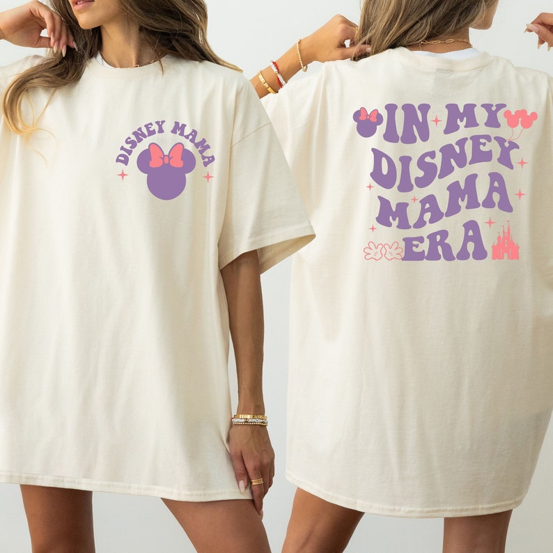 May include: A cream-coloured t-shirt featuring the text "DISNEY MAMA" and a Minnie Mouse graphic on the front. The back of the shirt displays the phrase "IN MY DISNEY MAMA ERA" in purple, accompanied by Disney-themed graphics, including a castle and balloons.