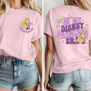 May include: Pink t-shirt with a graphic of Rapunzel on the front and the text "In My Disney Princess Era!" on the back. The shirt is being worn by a person with long blonde hair and denim shorts.
