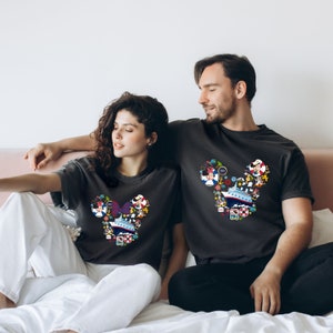 Pu&ograve; includere: Due magliette grigio scuro con un design a forma di testa di Topolino riempito di illustrazioni colorate a tema crociera. Il design include una nave da crociera, personaggi ed elementi nautici. Le magliette sono indossate da una coppia seduta su un letto.