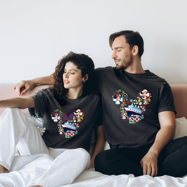 Pu&ograve; includere: Due magliette grigio scuro con un design a forma di testa di Topolino riempito di illustrazioni colorate a tema crociera. Il design include una nave da crociera, personaggi ed elementi nautici. Le magliette sono indossate da una coppia seduta su un letto.