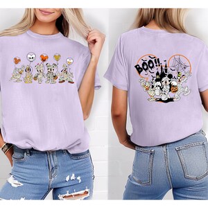 Puede incluir: Camiseta lila con un dise&ntilde;o de Halloween. La parte delantera muestra personajes de dibujos animados disfrazados de momias sosteniendo globos de calabaza. La parte trasera tiene la palabra "BOO!!" y una escena de Halloween.