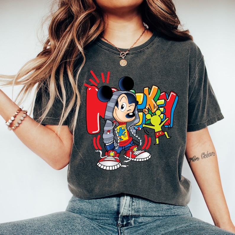 Pode incluir: Uma camiseta cinza-chumbo com um gr&aacute;fico colorido do Mickey Mouse vestindo um moletom cinza e t&ecirc;nis vermelhos. O gr&aacute;fico tamb&eacute;m inclui a palavra "Mickey" em letras coloridas.