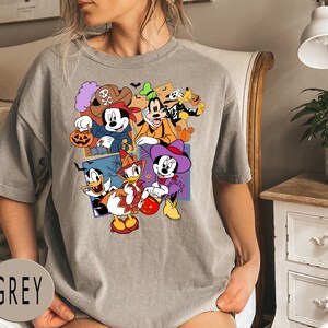 K&ouml;nnte beinhalten: Ein graues T-Shirt mit einem bunten Grafikdruck von Mickey Mouse und seinen Freunden in Halloween-Kost&uuml;men. Die Grafik zeigt Mickey Mouse, Minnie Mouse, Donald Duck, Goofy und Pluto.