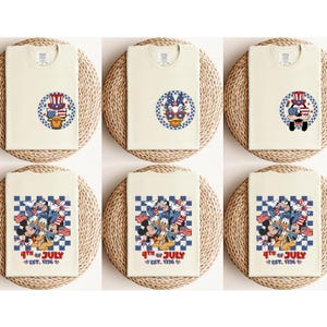 Pode incluir: T-shirts creme com desenhos de personagens de desenhos animados patri&oacute;ticos. Os desenhos incluem Pato Donald, Mickey Mouse e outros personagens usando chap&eacute;us e &oacute;culos de sol patri&oacute;ticos. O texto "4th of July EST. 1776" tamb&eacute;m est&aacute; presente.
