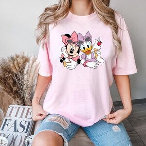 Puede incluir: Una camiseta rosa con un gr&aacute;fico de Minnie Mouse y Daisy Duck con gafas de sol y sosteniendo conos de helado. El gr&aacute;fico es de estilo de dibujos animados.