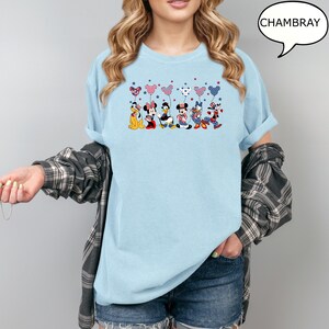 Puede incluir: Camiseta azul claro con un gr&aacute;fico de personajes de Disney sosteniendo globos en forma de coraz&oacute;n con estrellas y rayas patri&oacute;ticas. La palabra "CHAMBRAY" est&aacute; en un bocadillo. La camiseta es de estilo casual.