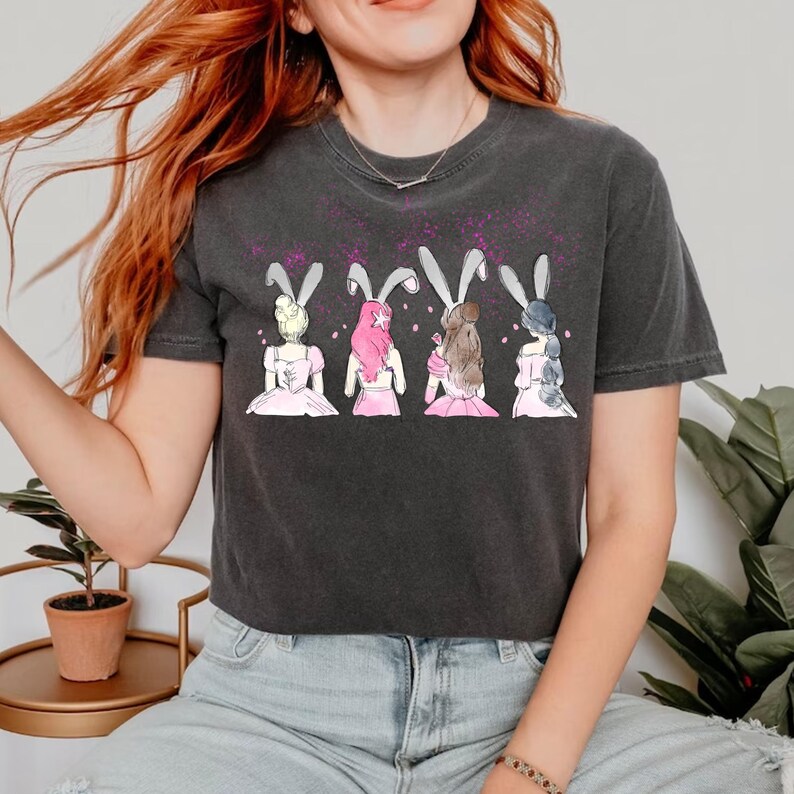 Puede incluir: Una camiseta gris oscuro recortada con un gr&aacute;fico de cuatro chicas con orejas de conejo que llevan vestidos rosas. Las chicas est&aacute;n de espaldas al espectador. El fondo es gris claro con un sutil patr&oacute;n moteado.