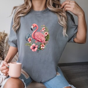 K&ouml;nnte beinhalten: Ein graues T-Shirt mit einem pinkfarbenen Flamingo und tropischen Blumen, die auf der Vorderseite aufgedruckt sind.