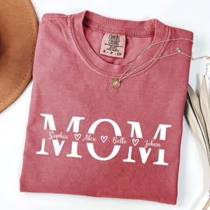 May include: A dusty rose Comfort Colors t-shirt with the word "MOM" in white block letters. Below the word "MOM" are the names Sophia, Alex, Belle, and Johan, each with a heart symbol. A silver necklace with a pendant is on the shirt.