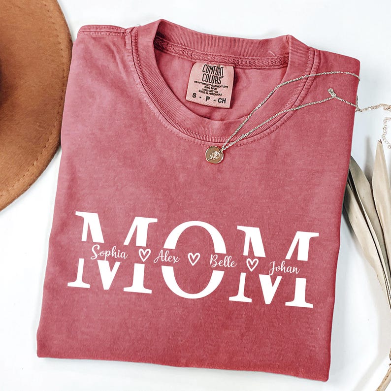 May include: A dusty rose Comfort Colors t-shirt with the word "MOM" in white block letters. Below the word "MOM" are the names Sophia, Alex, Belle, and Johan, each with a heart symbol. A silver necklace with a pendant is on the shirt.