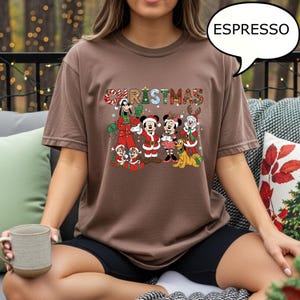 May include: A brown t-shirt with a Christmas-themed graphic. The design features Disney characters in festive outfits, with the word "CHRISTMAS" in colourful lettering. The shirt is a casual style, perfect for the holiday season.