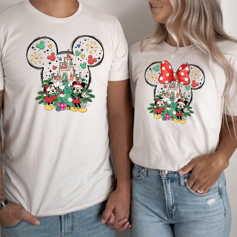 Puede incluir: Una camiseta blanca con un dise&ntilde;o de Mickey Mouse con tem&aacute;tica navide&ntilde;a que presenta a Mickey y Minnie Mouse de pie frente a un &aacute;rbol de Navidad y el castillo de Cenicienta. El dise&ntilde;o est&aacute; rodeado por un contorno negro de las orejas de Mickey Mouse.