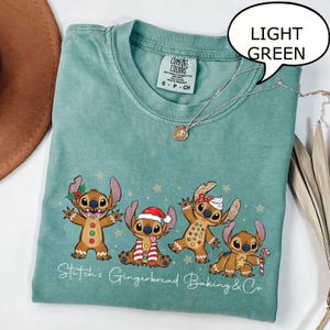 Puede incluir: Camiseta verde claro con cuello redondo y el texto "Stitch's Gingerbread Baking & Co." La camiseta presenta un gr&aacute;fico de cuatro personajes Stitch estilo pan de jengibre. Un collar dorado est&aacute; drapeado sobre la camiseta.