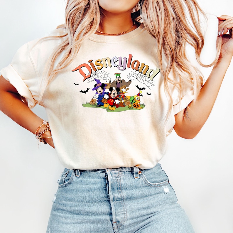 Puede incluir: Una camiseta blanca con un logotipo de Disneyland de estilo vintage y un gr&aacute;fico de tem&aacute;tica de Halloween con Mickey Mouse, Donald Duck, Goofy y Pluto vestidos con disfraces. El gr&aacute;fico incluye murci&eacute;lagos, telara&ntilde;as y calabazas.