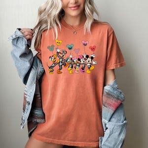 May include: A burnt orange t-shirt with a graphic of Mickey Mouse, Minnie Mouse, Pluto, and Goofy dressed as skeletons holding colorful balloons. The characters are all smiling and appear to be walking in a line.