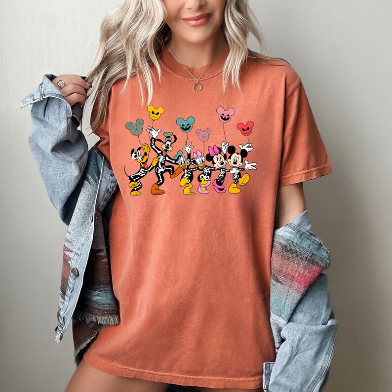 May include: A burnt orange t-shirt with a graphic of Mickey Mouse, Minnie Mouse, Pluto, and Goofy dressed as skeletons holding colorful balloons. The characters are all smiling and appear to be walking in a line.