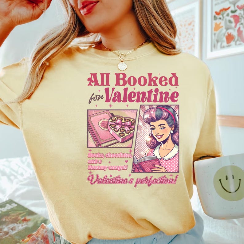 Puede incluir: Una camiseta amarilla con el texto "All Booked for Valentine" en rosa y blanco. La camiseta presenta una ilustraci&oacute;n de dibujos animados de una mujer leyendo un libro con una caja de chocolates y un libro en el fondo. El texto debajo de la ilustraci&oacute;n dice "Books, chocolates, and a dreamy escape! Valentine's perfection!"
