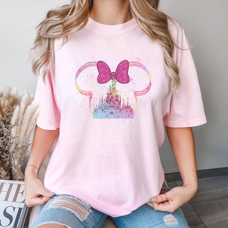 Puede incluir: Camiseta rosa con un dise&ntilde;o de acuarela de un castillo con un lazo rosa en la parte superior. El castillo est&aacute; rodeado de un dise&ntilde;o de acuarela rosa, azul y amarillo.