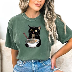 Puede incluir: Una camiseta verde con un gato negro comiendo fideos ramen de un cuenco blanco. El gato sostiene palillos en sus patas.