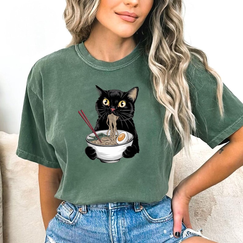 Puede incluir: Una camiseta verde con un gato negro comiendo fideos ramen de un cuenco blanco. El gato sostiene palillos en sus patas.