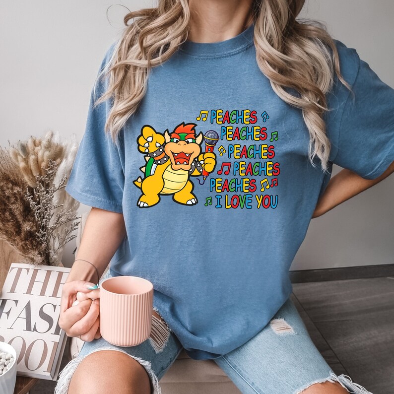 May include: A blue t-shirt features a colorful graphic of a cartoon character holding a microphone, with the words "PEACHES" and "I LOVE YOU" in a playful font. The character is yellow, orange, and green.